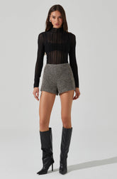 A woman is wearing a black, long-sleeved, semi-sheer, high-neck top with vertical stripes. She pairs it with gray, high-waisted, tailored shorts with a textured fabric. She completes the outfit with knee-high black leather boots with pointed toes and stiletto heels.